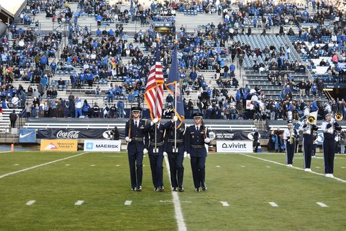 4 men dressed in military holding rifles and the American flag and blue flag in a ceremonious formation, standing on a football field