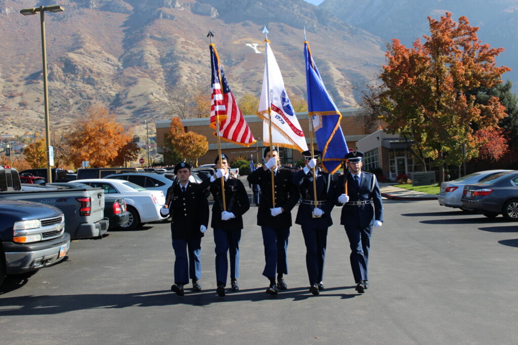 5 men dressed in military holding rifles and the American flag and two other flags in a ceremonious formation, walking in a BYU parking lot.