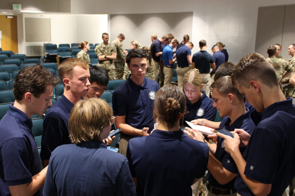 Group of men and women standing in a circle talking. They are wearing tan khakis and blue polos.