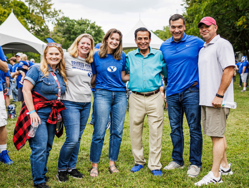 Six people smile together on a grassy lawn at an outdoor event.