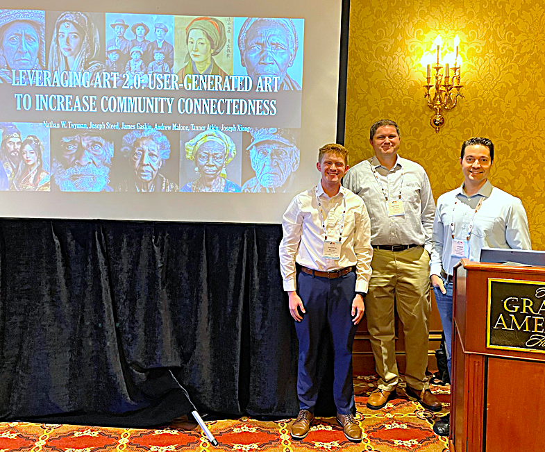 One male students stand with two male faculty members next to a large presentation screen