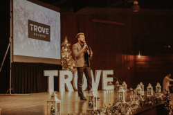 Blake Marchant, a white man with red hair and facial hair, giving a speech on a stage with the word Trove in the background.