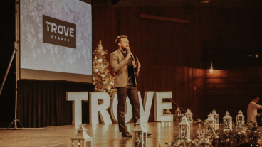 Blake Marchant, a white man with red hair and facial hair, giving a speech on a stage with the word Trove in the background.