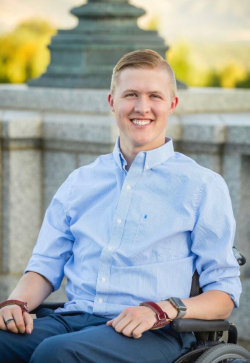 Josh Hinton, a white male wearing a blue button-down shirt. He is sitting in a wheelchair and smiling.