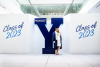 Woman in regalia standing in front of a giant blue Y with Class of 2023 written on wall