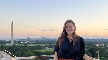 Lulu Gilbert standing on the roof of a building at sunset, smiling at the camera. The Washington Monument is in the skyline behind her.