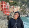 Tehani Travis smiles at the camera. Behind her is a city in Italy where a cliff falls abruptly into ocean, with brightly colored buildings reaching all the way to the edge.