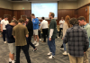 Students stand conversing in small groups, looking down at their phones. Several students are in the process of moving from one group to another. In the background a projected slide reads: "A Crisis Occurs!!!"