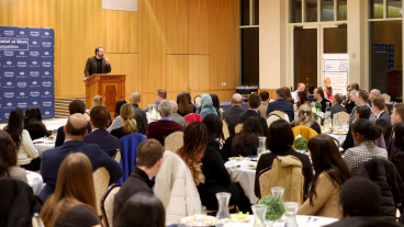 Man standing at a podium giving a speech in a room full of people sitting at dining tables.