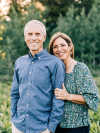 Mark and Karen Zimbelman stand in a field during golden hour with trees in the background, smiling widely at the camera.