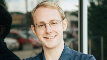 Aaron Adams leans against a window, smiling at the camera with his arms folded. He's wearing a BYU Marriott polo shirt and you can see the sky reflected in the window behind him.