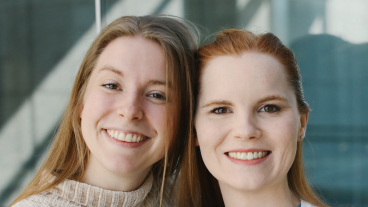 A close up of two young woman smiling at the camera. They both have long hair that is tucked back and are standing in front of a glass wall.