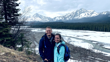 A man and a women dressed in hiking gear pose in the grass with a frozen river and snowy mountains in the background.