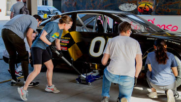 A group of five work on a retired racing car. In the center, a young woman is working the car-jack, her braid suspended in mid-air from the quick movement.