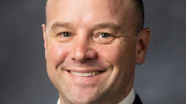 Richard Gardner smiles at the camera in a professional headshot with a gray background.