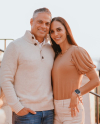 Tyler and Jerriann Banks stand together in the soft golden glow just before sunset. They're both smiling at the camera with one hand in their pockets and the other around each other.