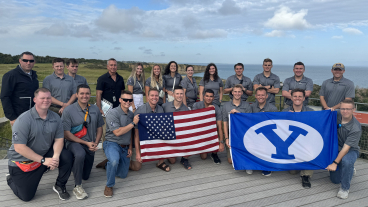 A group of student in faculty pose at the Pointe Du Hoc, holding both the US flag and a BYU flag.