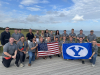 A group of student in faculty pose at the Pointe Du Hoc, holding both the US flag and a BYU flag.