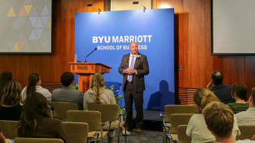 Jay Meldrum wears suit and boutonnière and stands in front of podium with a blue BYU Marriott backdrop