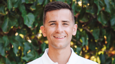 Adam Boswell wears white polo and smiles in front of green foliage.