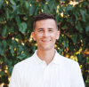 Adam Boswell wears white polo and smiles in front of green foliage.