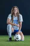 Dressed in a BYU athletic uniform, Folino smiles at the camera as she kneels on a soccer field with a soccer ball.