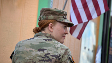 Erin Cole, in army uniform, looks to her right with an American flag draping in the background.