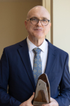 Robert Carver smiles in a suit while holding a wooden award from BYU MPA.