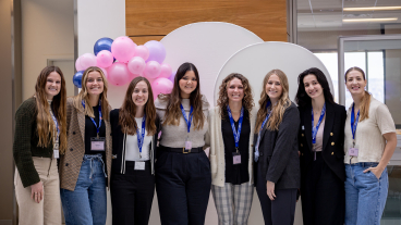 A group of female attendees smile at the camera, wearing blue lanyards and with pink balloons in the background.