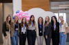 A group of female attendees smile at the camera, wearing blue lanyards and with pink balloons in the background.