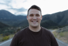 A man with brown hair wearing a brown short sleeve shirt smiles for a photo with a paved road and mountains behind him.