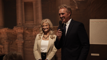 A man wearing a navy blue suit and a woman wearing a tan blazer stand smiling toward a crowd with a beige and brown backdrop. The man is holding a black microphone near his mouth.