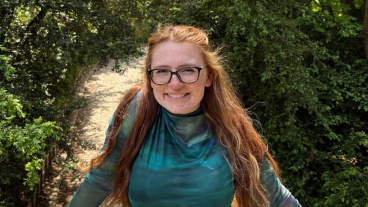 Sarah-Jane Tate stands on a bridge over a path surrounded by trees, smiling at the camera.