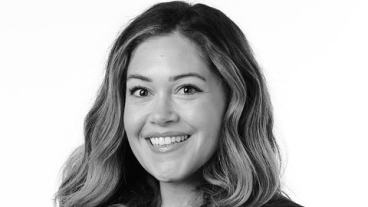 In black-and-white; a woman wearing a black blazer and top smiles for a professional headshot with her arms crossed.