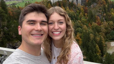 A college-aged couple smiles for a selfie in front of green, yellow, and orange trees with the Neuschwanstein castle in the background.
