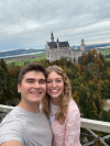 A college-aged couple smiles for a selfie in front of green, yellow, and orange trees with the Neuschwanstein castle in the background.