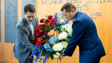 Two men place an easel with a red, white, and blue flower wreath in front of the room. One man is wearing a grey suit and the other is wearing a navy blue formal military suit.