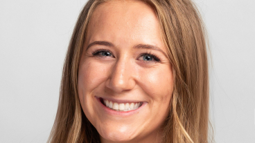 A blonde young woman in a white button-down shirt and a black blazer smiles for a professional headshot.