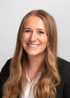 A blonde young woman in a white button-down shirt and a black blazer smiles for a professional headshot.