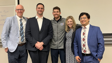 A group of four professionally dressed adults smile for a group picture with a man in a Chips Ahoy jacket in a classroom.
