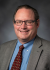 Tyler Shumway, wearing glasses and a gray suit, blue dress shirt, and red tie smiles for a professional headshot.