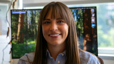 A woman in a blue and white striped button-down shirt smiles for a picture in front of a computer monitor.
