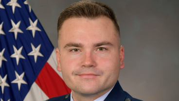 A man with brown hair and brown eyes smiles while wearing a navy blue air force uniform. The uniform has colorful badges over his right chest.