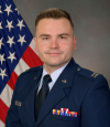 A man with brown hair and brown eyes smiles while wearing a navy blue air force uniform. The uniform has colorful badges over his right chest.