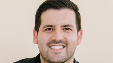 Andres Aleson smiles for a professional headshot wearing a black denim jacket over a dark-red T-shirt.