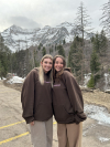 Two young adult women wearing light brown pants and dark brown hoodies stand smiling in front of the mountains.