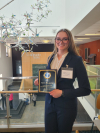 A young adult woman with light hair and glasses wears a navy blue blazer and holds a plaque that reads “1st Place.”