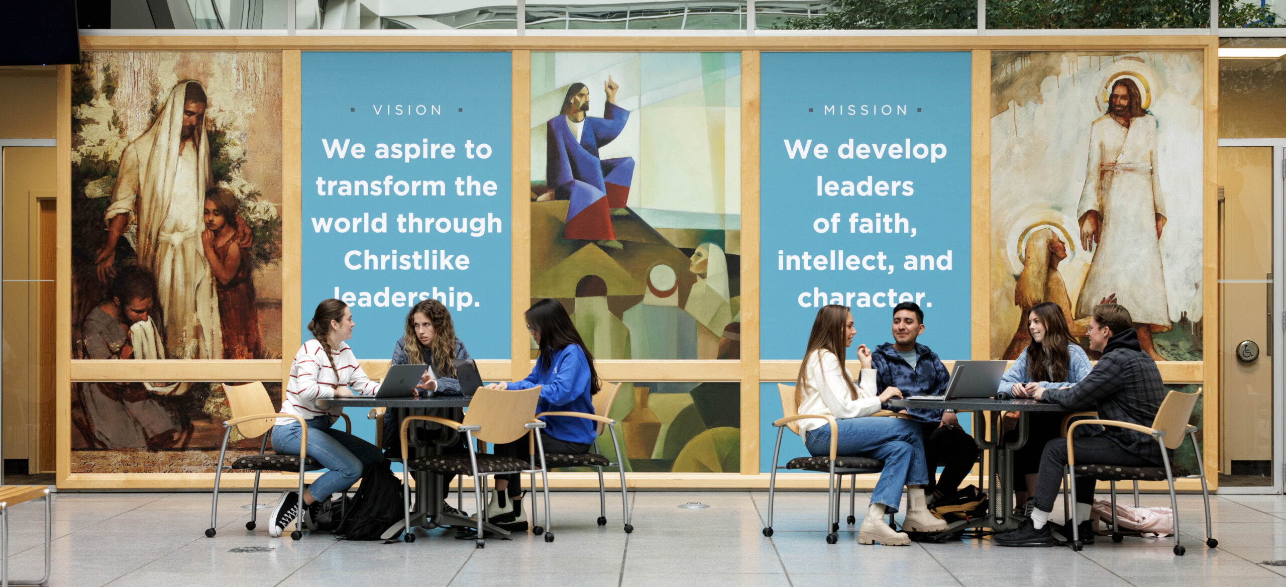 Wall with photo of Christ and BYU Marriott mission and vision. Students sitting at tables in front of wall.